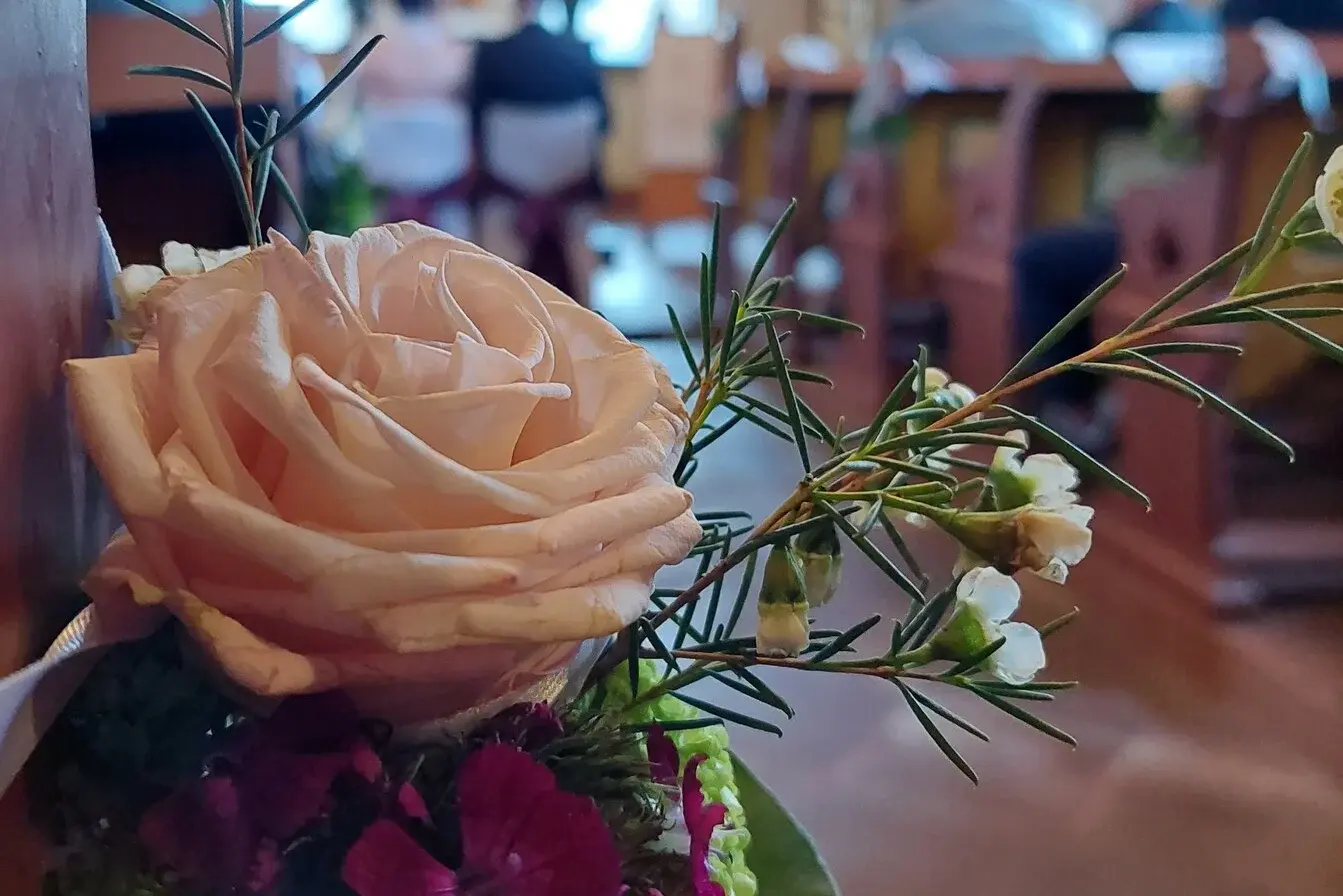 Ein Rosengebinde schmückt die Kirchenbank während einer kirchlichen Trauung. Im Hintergrund sitzt ein Paar vor dem Altar. Die Blume steht im Fokus, der Hintergrund ist unscharf.