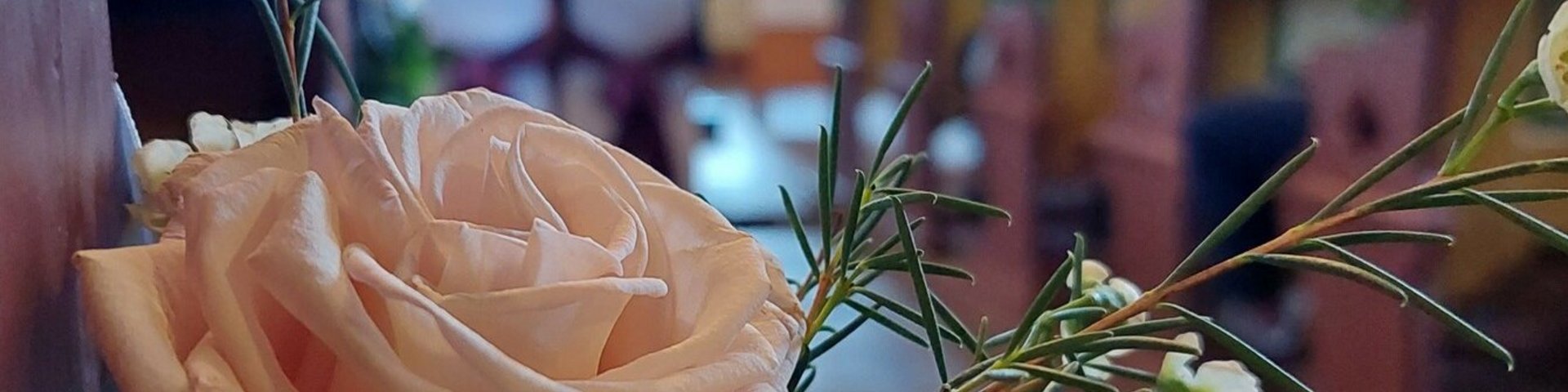 Ein Rosengebinde schmückt die Kirchenbank während einer kirchlichen Trauung. Im Hintergrund sitzt ein Paar vor dem Altar. Die Blume steht im Fokus, der Hintergrund ist unscharf.